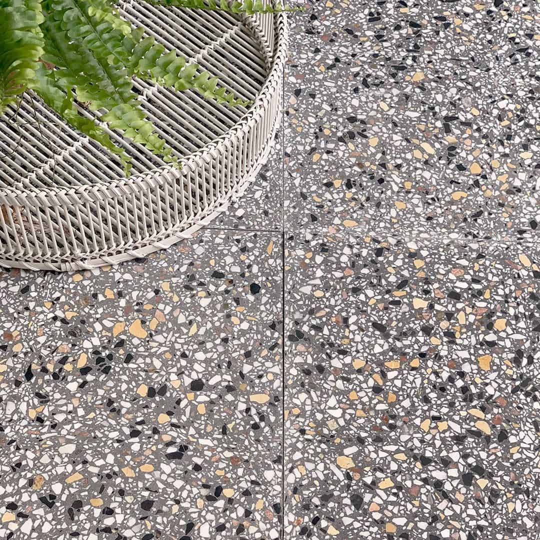 Gray terrazzo tile flooring with multicolor stone chips, paired with a woven round planter and green fern leaves.