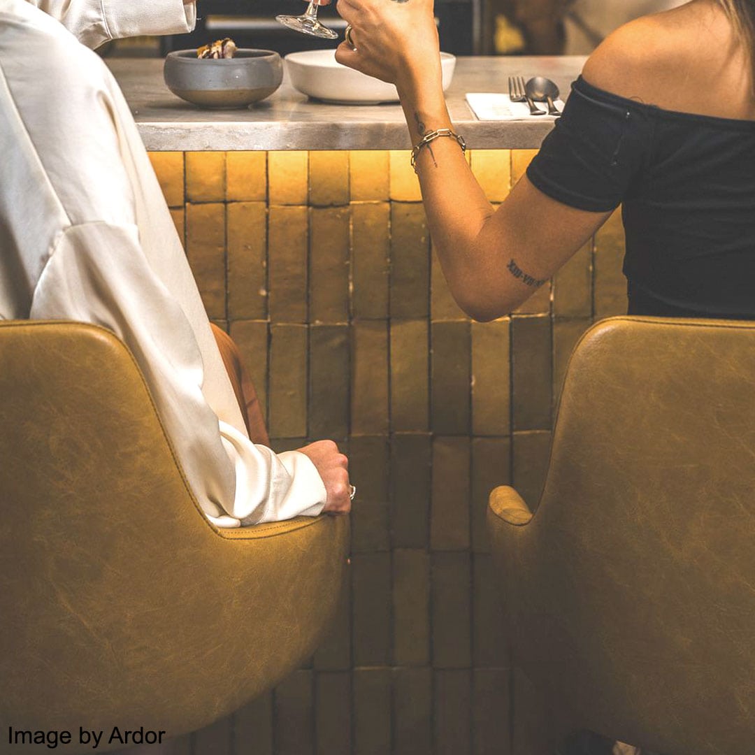 Elegant bar counter with polished marble surface and golden textured front, set with wine glasses, cutlery, napkins, and greenery vases, illuminated by metallic pendant lights in a stylish restaurant.