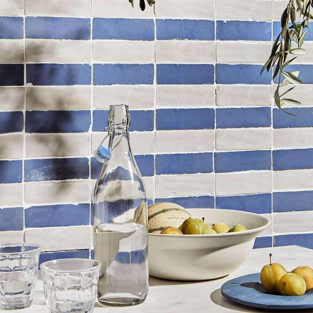 Table against blue-and-white tiled wall with glass bottle of water, two empty glasses, white bowl of fruit, blue plate with plums, and olive branches casting shadows.
