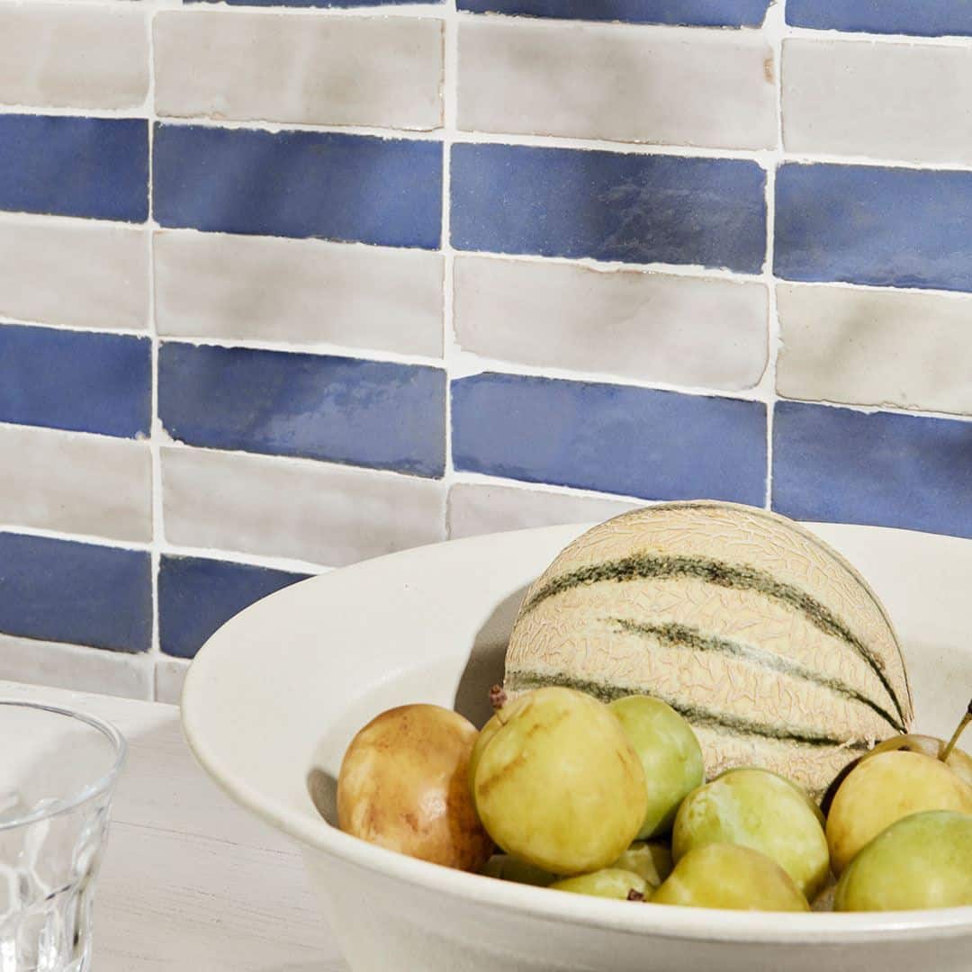 Table against blue-and-white tiled wall with glass bottle of water, two empty glasses, white bowl of fruit, blue plate with plums, and olive branches casting shadows.