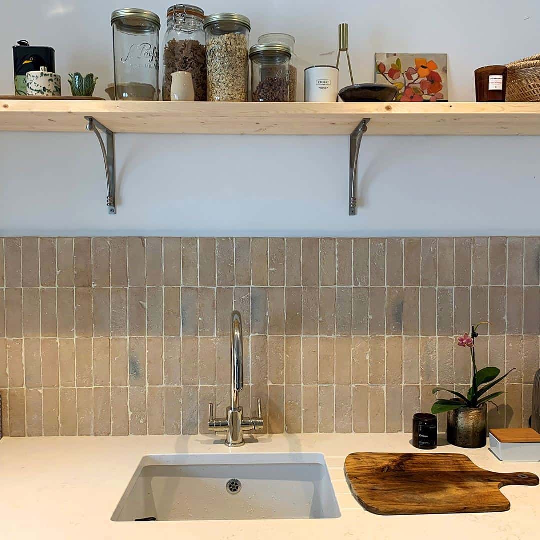 Modern kitchen with a minimalist design, featuring beige tiled backsplash, a sink, wooden cabinetry, and an open shelf with jars and decor items.