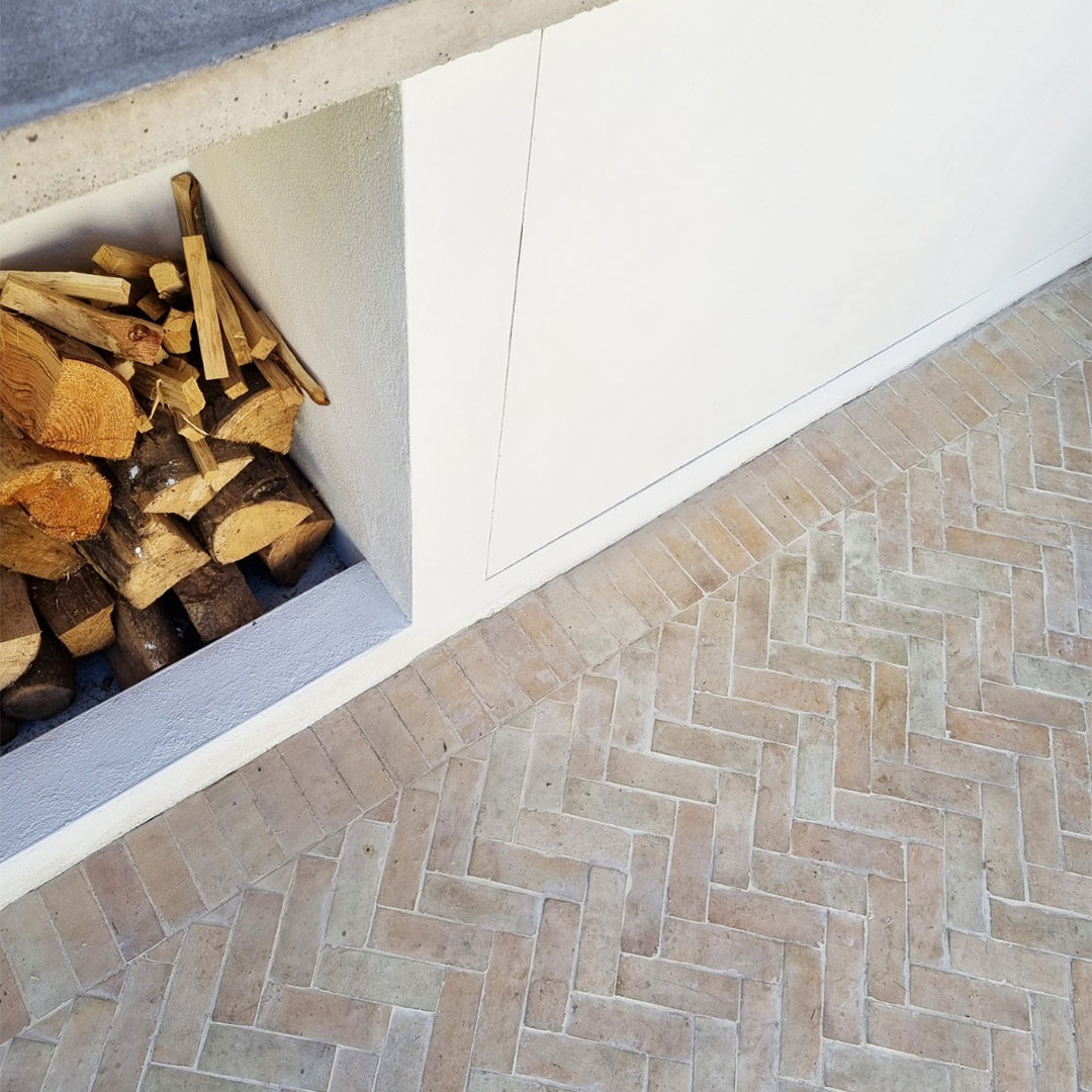 Stack of firewood next to a white wall with a herringbone stone floor.