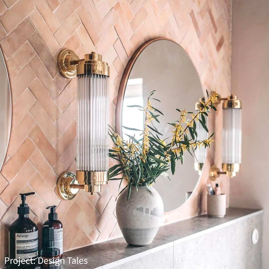 Elegant bathroom featuring a pink herringbone tile wall, round mirrors, gold light fixtures, and a vase with plants. A few luxury bottles are placed on the countertop.