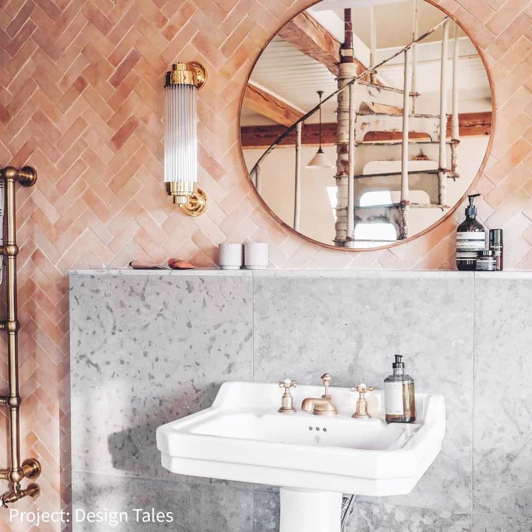 Stylish bathroom with a pink herringbone tile wall, a round mirror, vintage light fixtures, and a stone countertop with luxury bottles. A spiral staircase is reflected in the mirror.