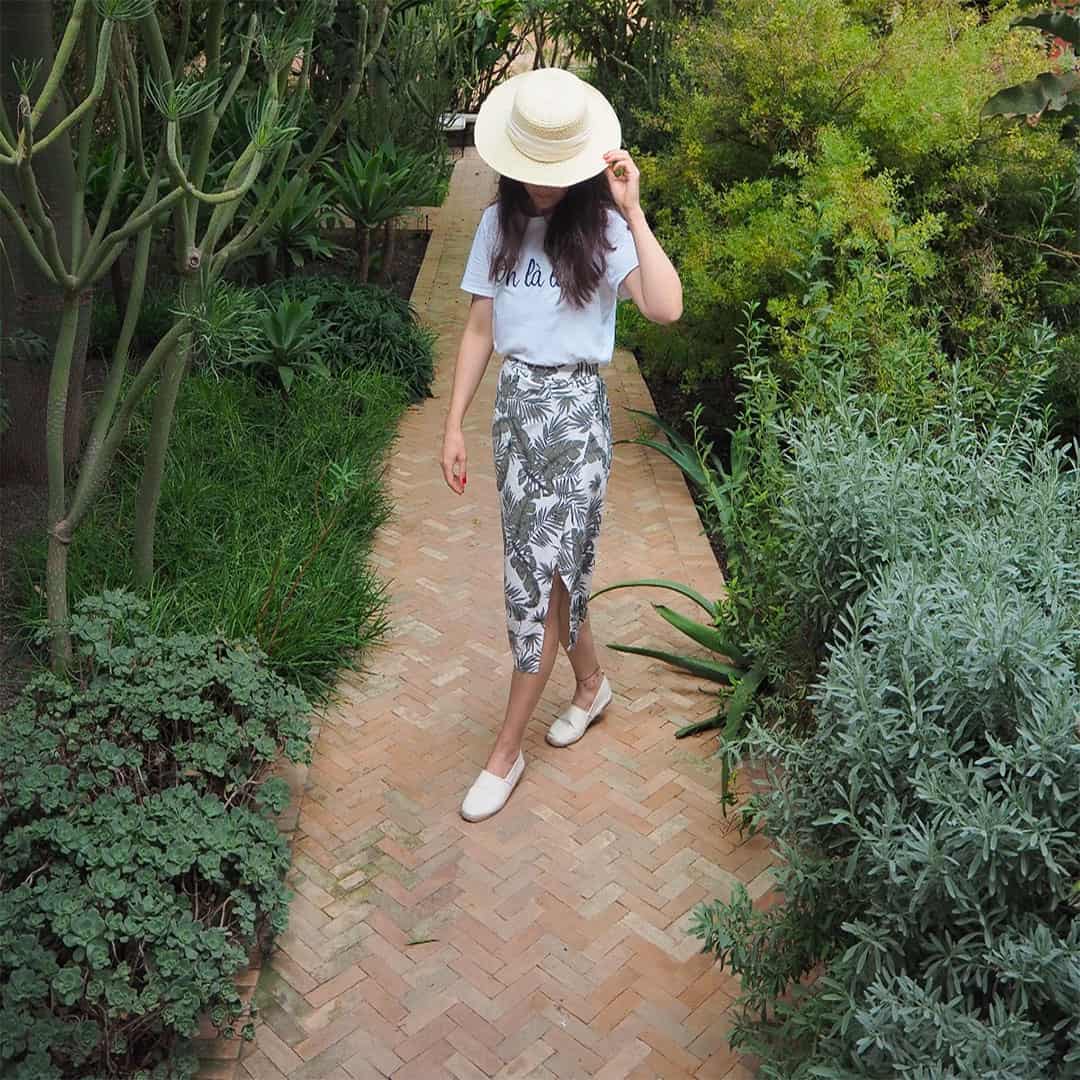 Woman walking through a lush garden path wearing a straw hat, a casual t-shirt, and a floral skirt.