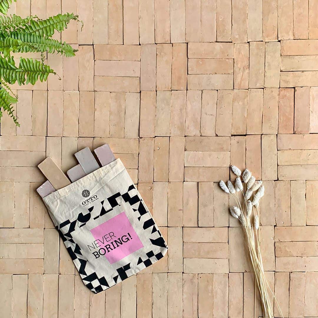 Stylish flat lay featuring a patterned tote bag with the text "NEVER BORING!" on a terracotta tile floor, accompanied by a fern and dried flowers.