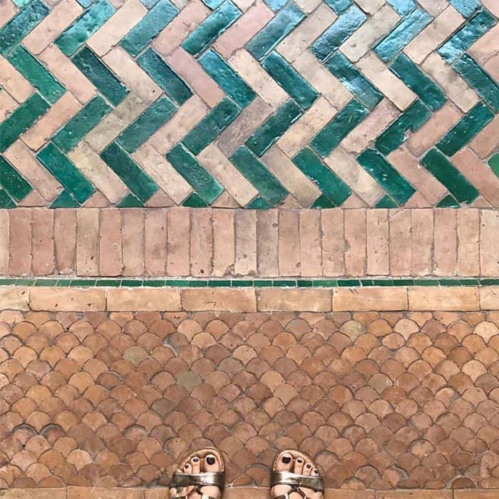 Close-up of a terracotta floor with herringbone green tiles, and a scalloped pattern below, with a person’s sandals in the frame.