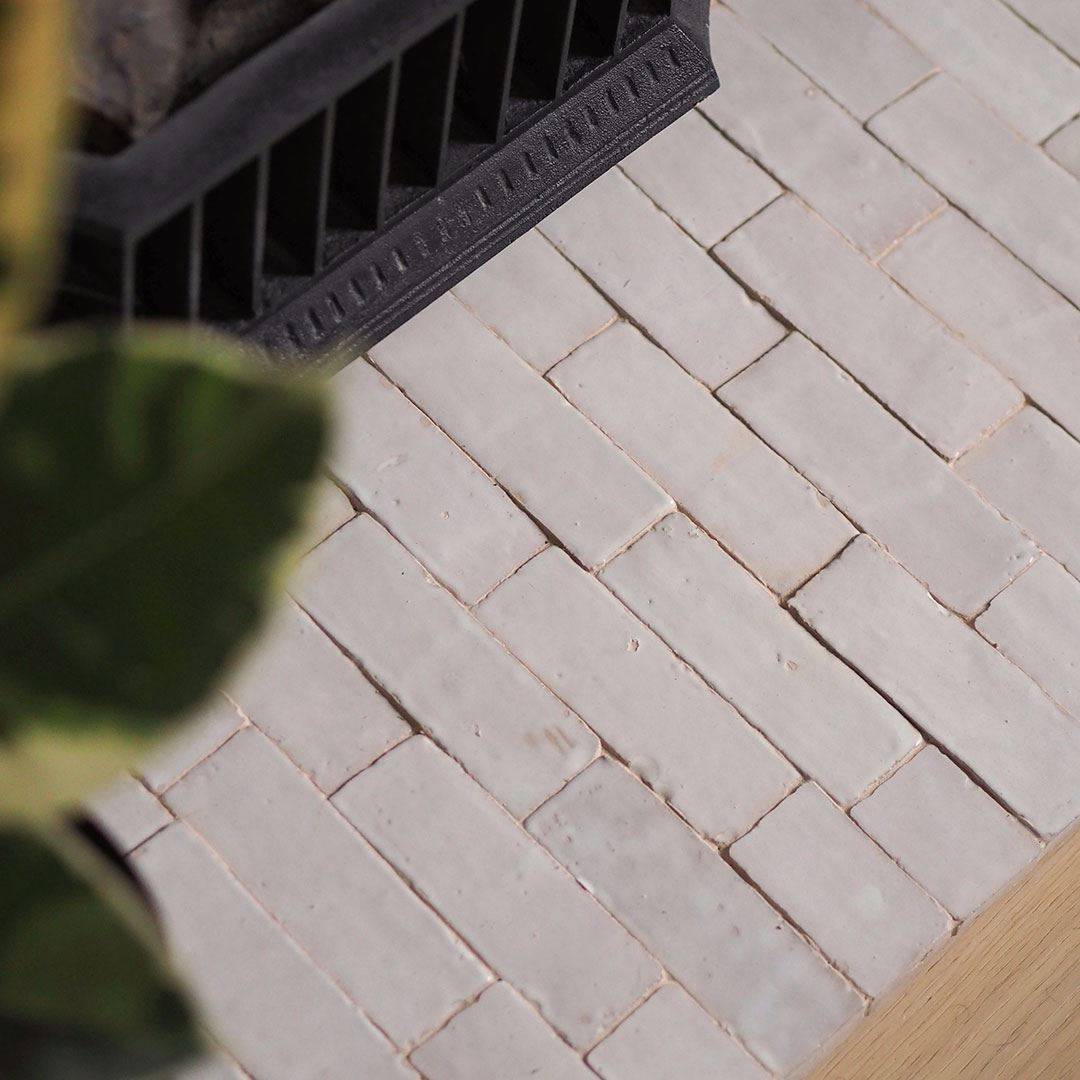 Close-up of a light beige rectangular tile surface arranged in a staggered brick pattern, with part of a black metal grate and a green leafy plant visible in the corners.