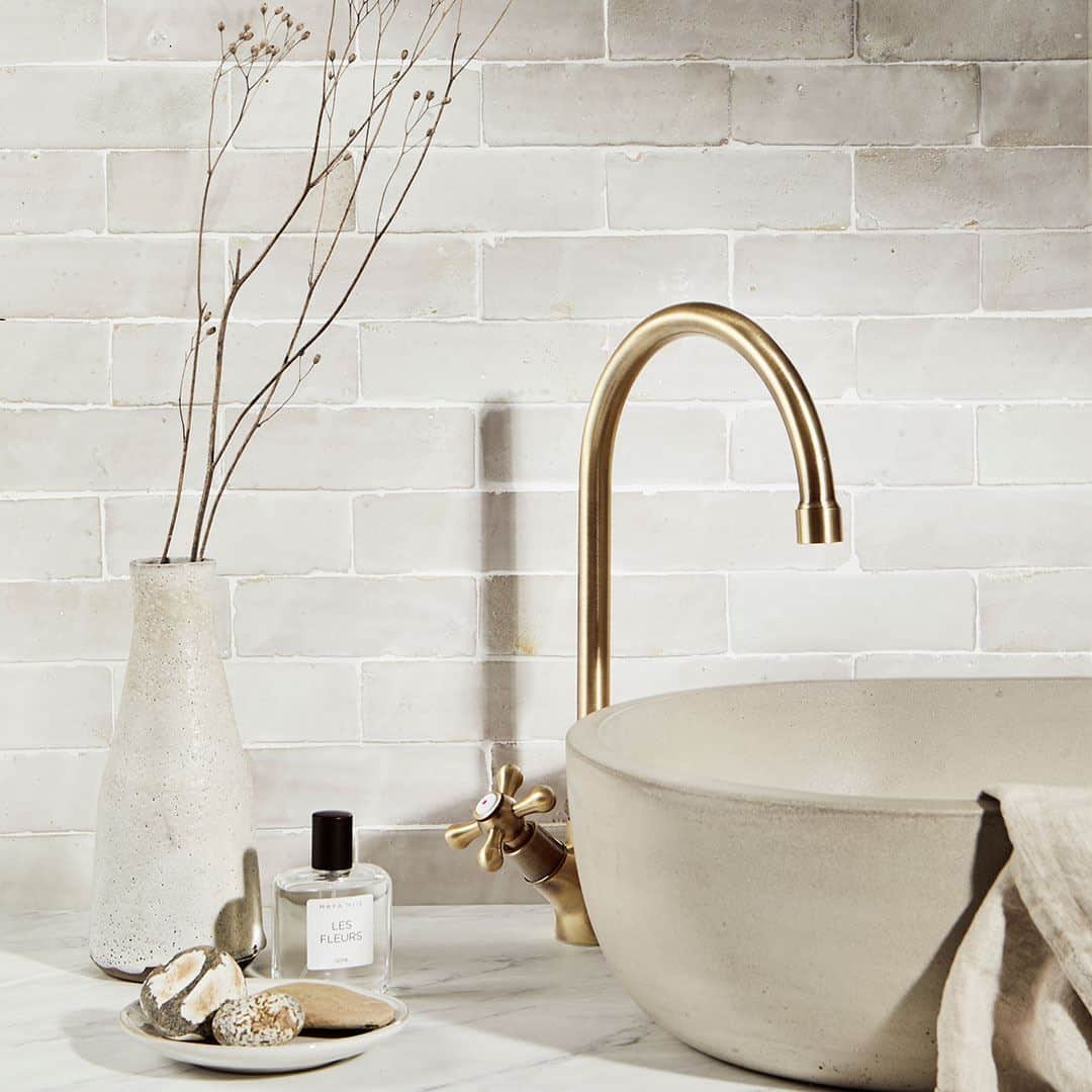 Minimalist bathroom sink setup featuring a round stone basin with a brushed brass faucet, a small vase with dried branches, a bottle of perfume labeled “Les Fleurs,” and a dish with natural stones, all set against a light beige tiled wall.