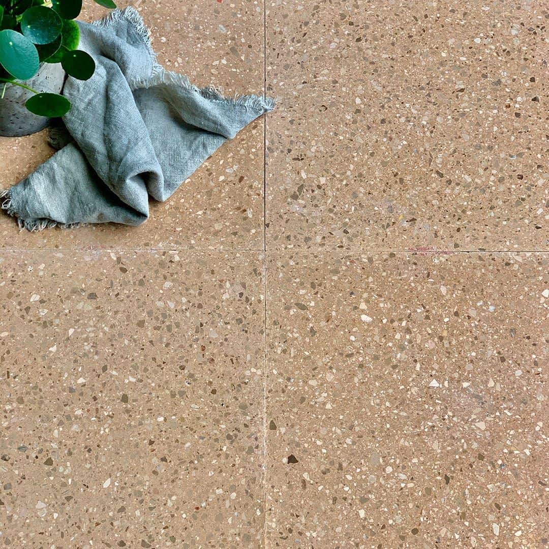Warm beige terrazzo tile floor with small stone chips, styled with a folded linen cloth and green plant in the corner.
