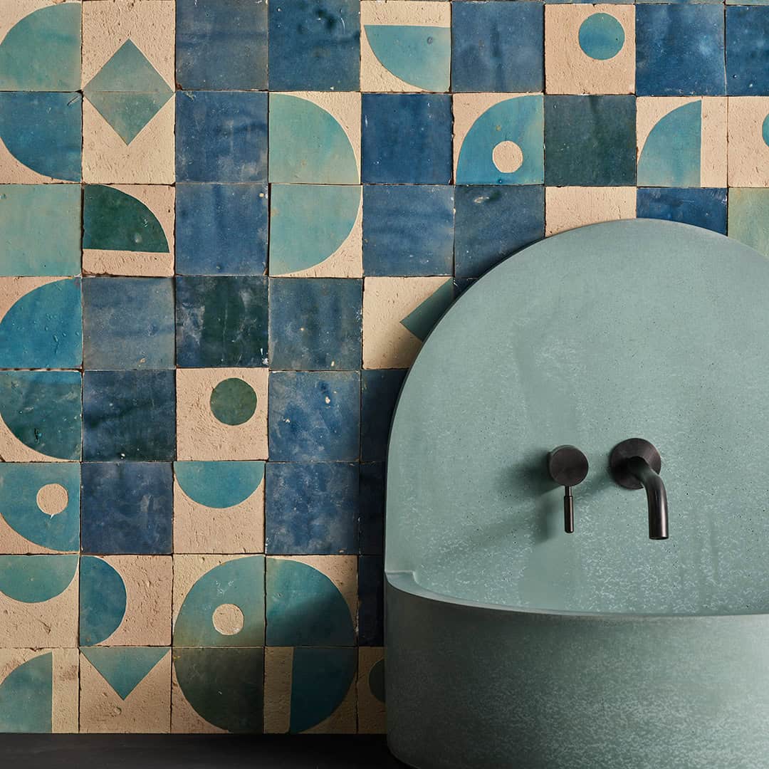 Close-up of a bathroom wall covered in blue and beige geometric patterned tiles featuring circles, semicircles, and triangles. In front of the wall is a light teal arched sink with a matte black wall-mounted faucet.