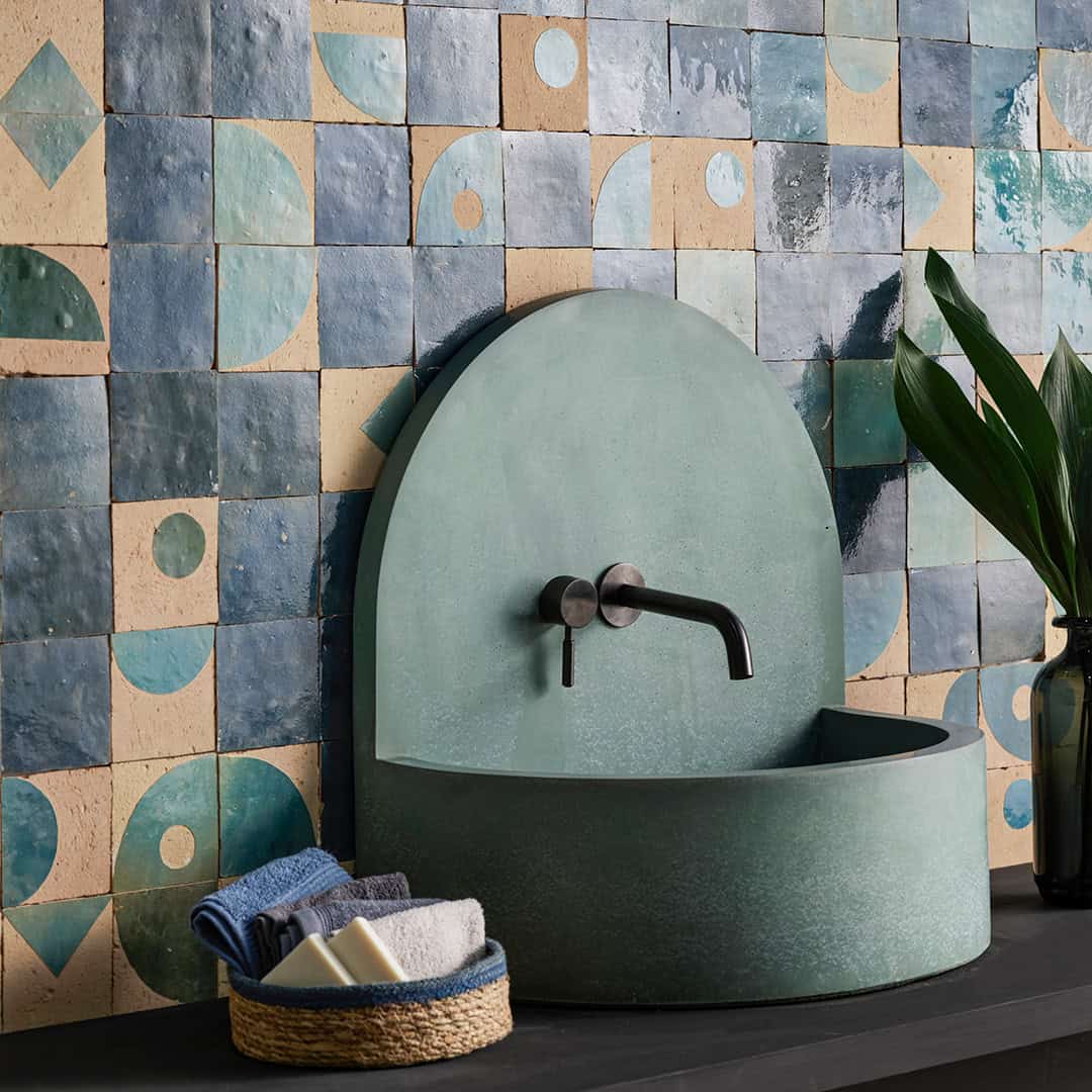 Wall tiles in blue and beige geometric patterned tiles, and a black vase with green leaves sits beside the sink.