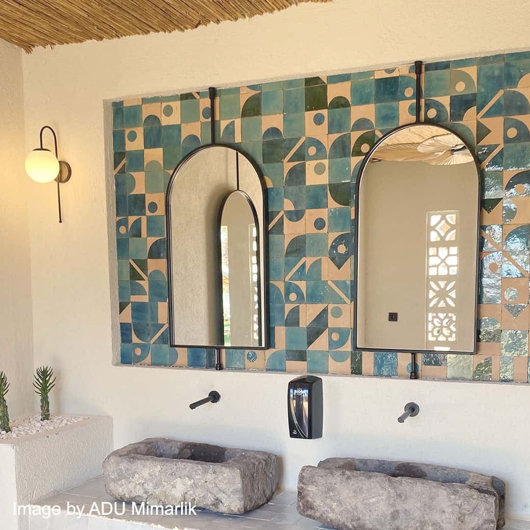 Bathroom with two stone sinks and wall-mounted black faucets beneath arched mirrors framed by blue and beige geometric patterned tiles.
