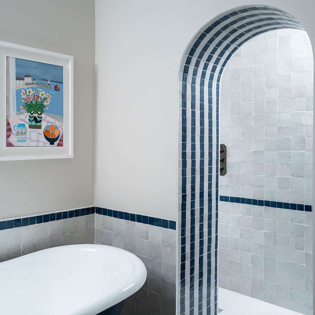 Modern bathroom with white bathtub, arched shower entrance framed by gray tiles with blue accents, and colorful still life artwork on the wall.