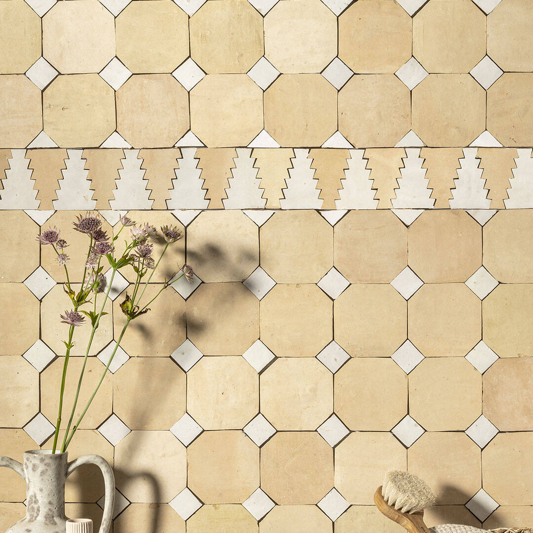 A wall with off-white tiles, zigzag border, and wooden brush and a vase with flower.