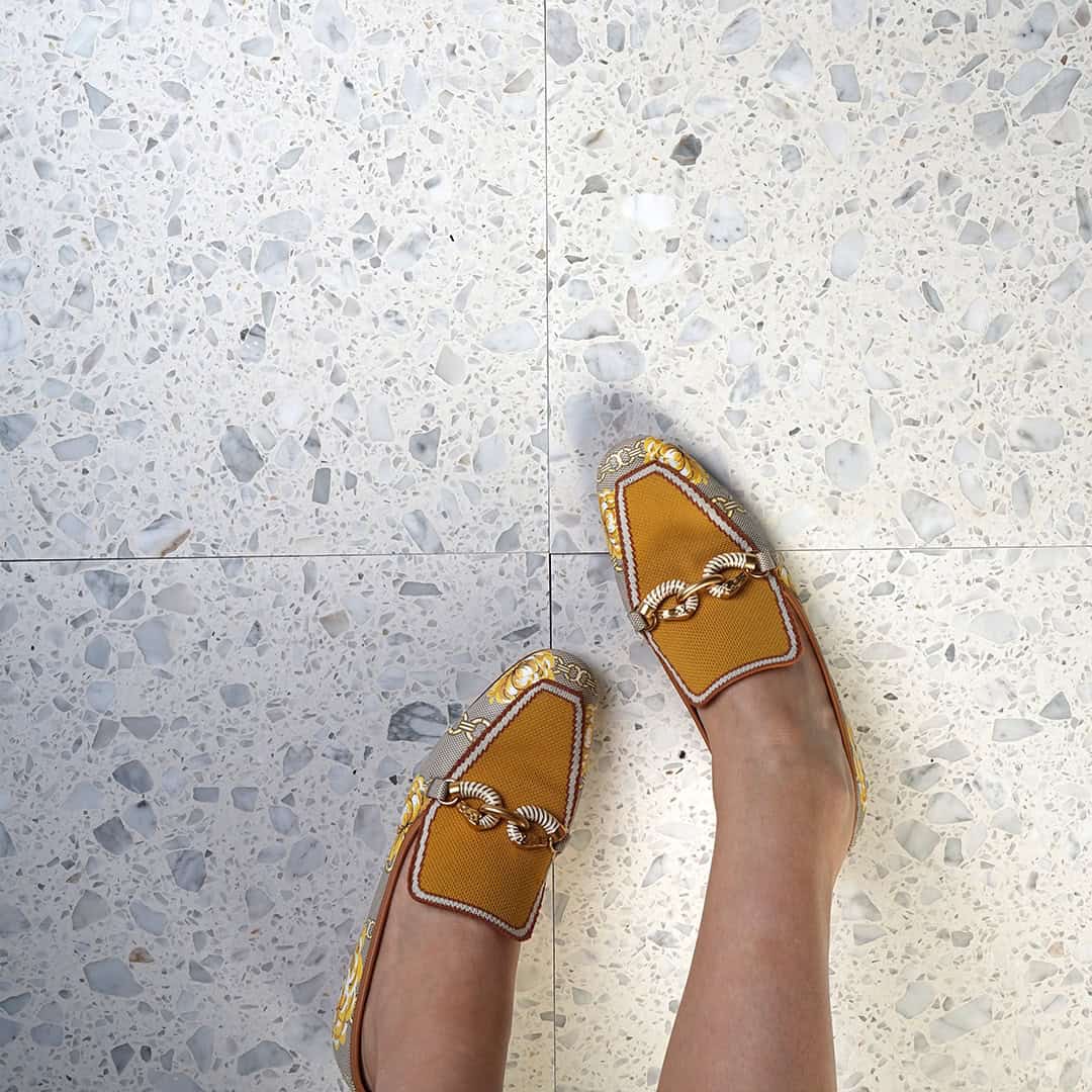 Person wearing mustard loafers standing on light terrazzo tile floor with subtle gray stone fragments.