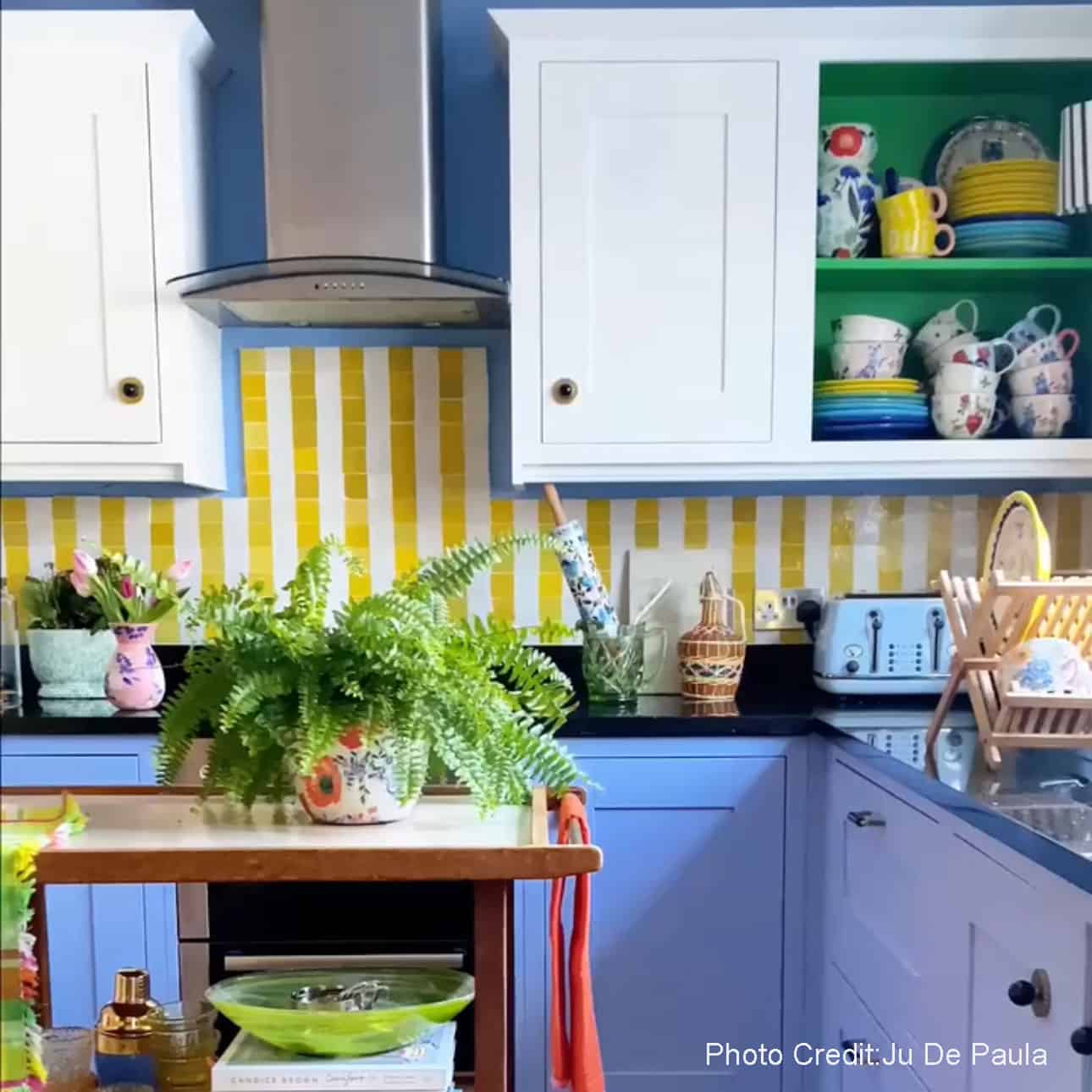 Colorful kitchen with blue cabinets, open shelving, and yellow striped tile backsplash, styled with plants and dishware.