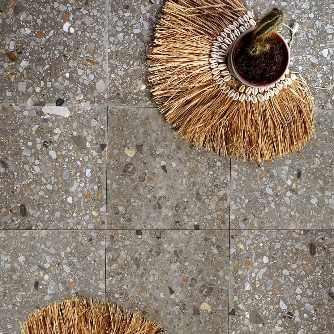 Neutral gray terrazzo tile floor with mixed stone chips, styled with woven mat and small cactus plant.