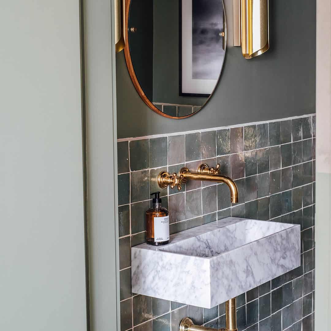 Elegant bathroom featuring green tiles, a marble sink, and brass faucet with a round mirror above.