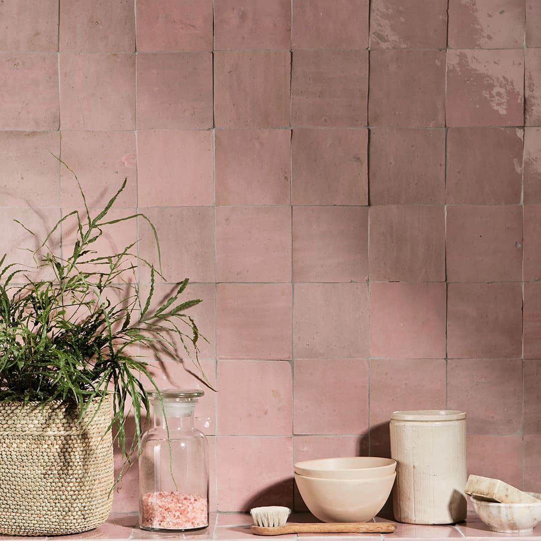 A countertop with pink square-tiled wall backdrop, featuring a woven basket holding a leafy green plant, a glass jar of pink salt, a wooden brush, a ceramic bowl, a cylindrical ceramic container, and two stacked smaller ceramic bowls, arranged neatly in a soft, natural aesthetic.