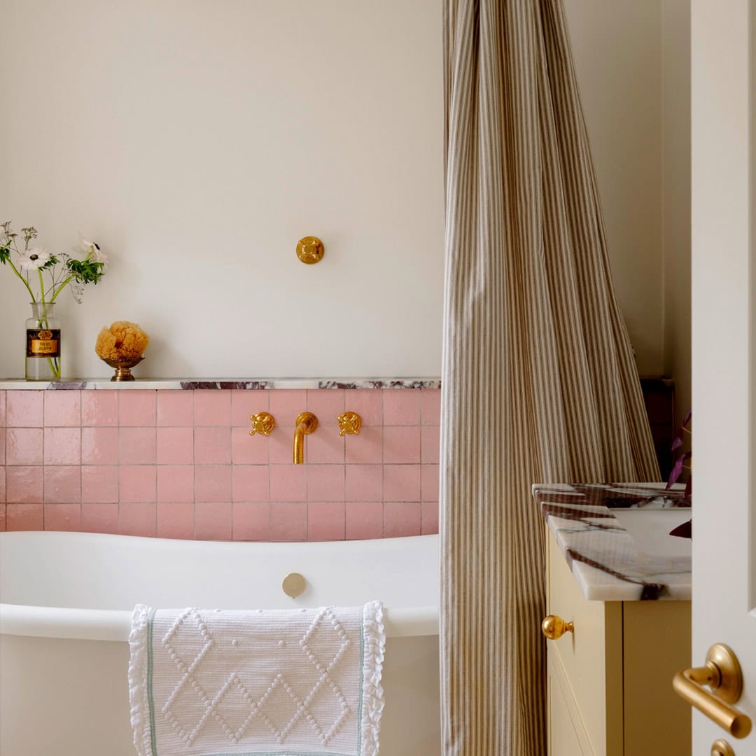 Glossy pink tiled wall topped by a marble ledge, gold faucet fixtures, a beige striped shower curtain, and decorative accents including flowers.