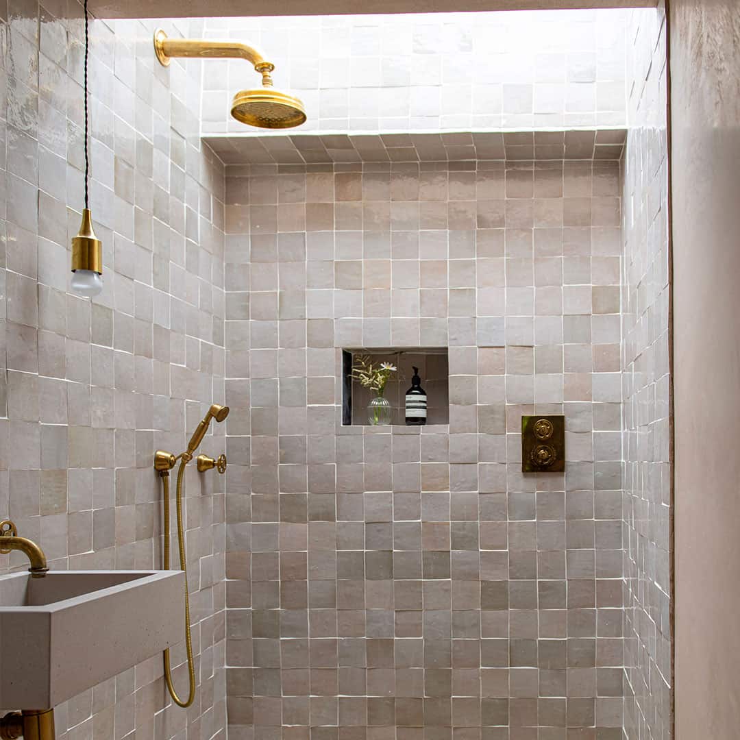 A modern shower area with beige square tiled walls and brass fixtures, including an overhead shower, handheld shower, and faucet.