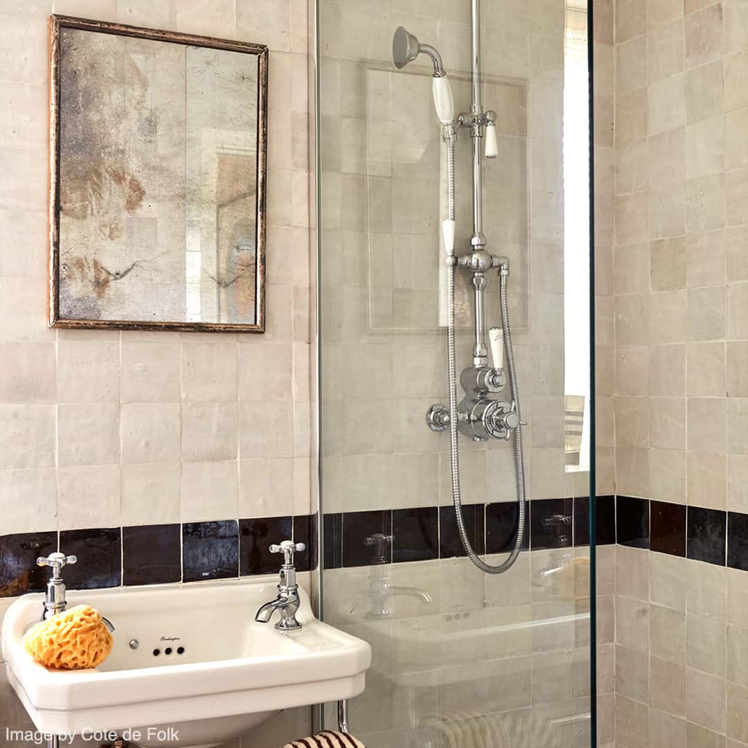 A modern shower area with beige square tiled walls and brass fixtures, including an overhead shower, handheld shower, and faucet.