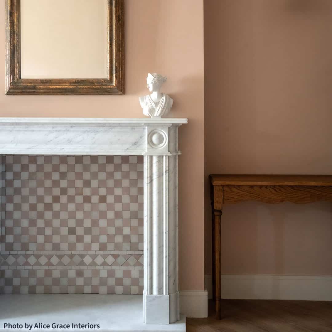 Elegant marble fireplace with checkered hearth, gold-framed mirror, bust sculpture, and peach-colored walls.