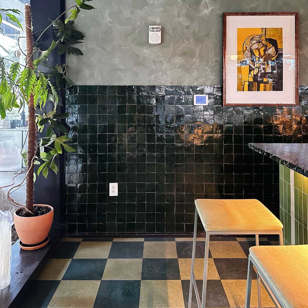 Modern interior with dark green glossy tiles, a black and yellow checkered floor, a framed abstract painting, and a plant in a pot.