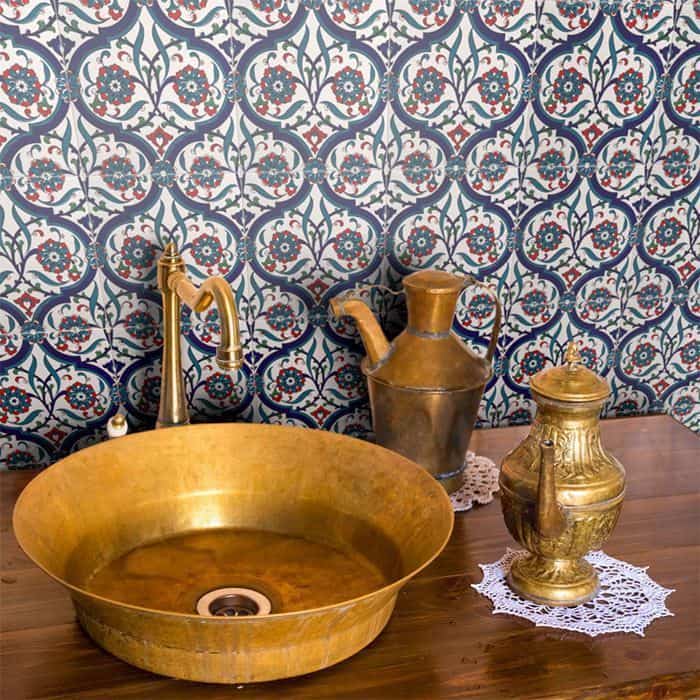 Decorative bathroom with brass basin and faucet, ornate blue-red patterned tile wall, wooden counter holding metallic vessels on lace doilies.