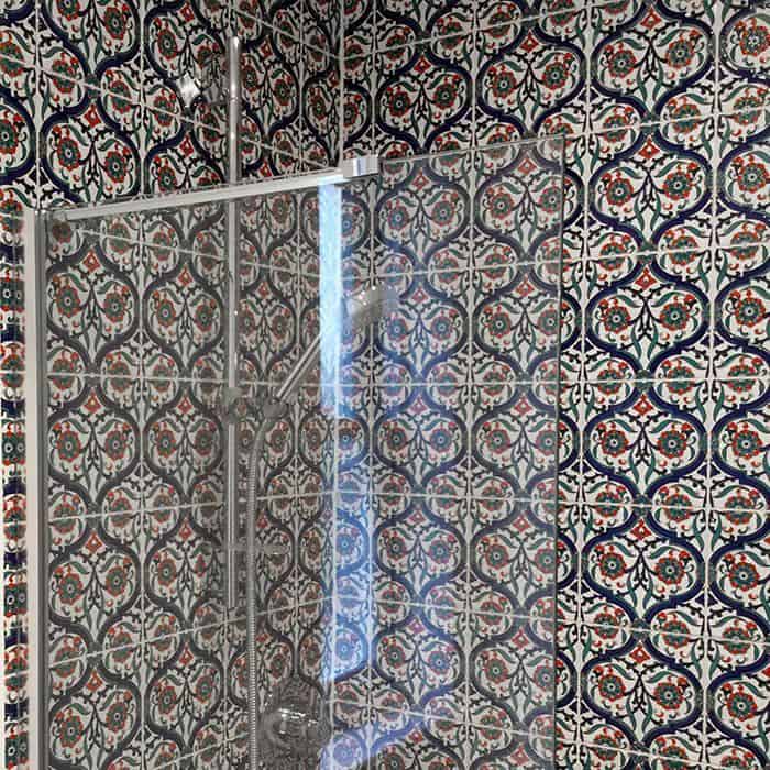 Shower with glass partition and ornate ceramic tile walls featuring blue, red, green, and white floral geometric patterns, with handheld showerhead on vertical bar.