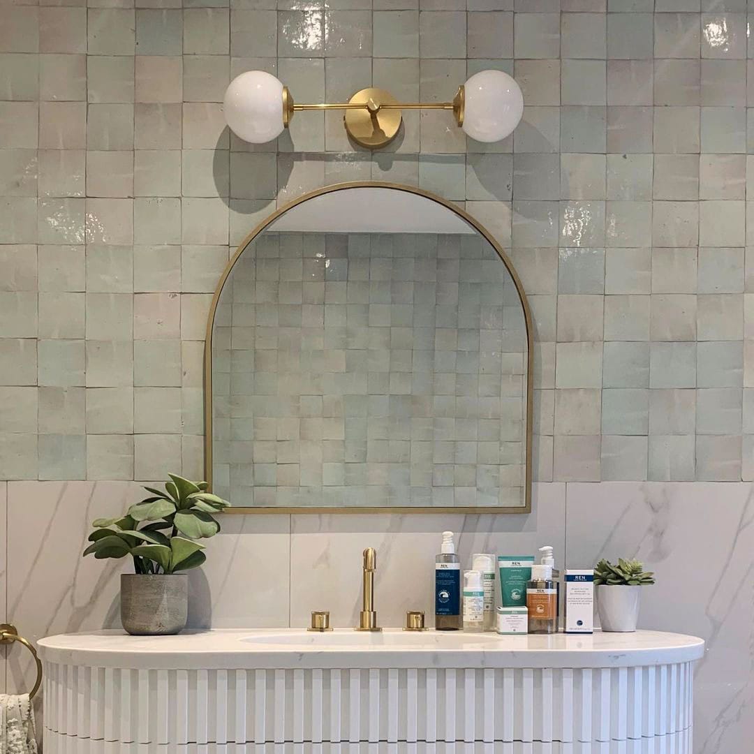 Elegant bathroom featuring a light green tiled wall, a gold-framed arch mirror, and a marble vanity with skincare products and plants.