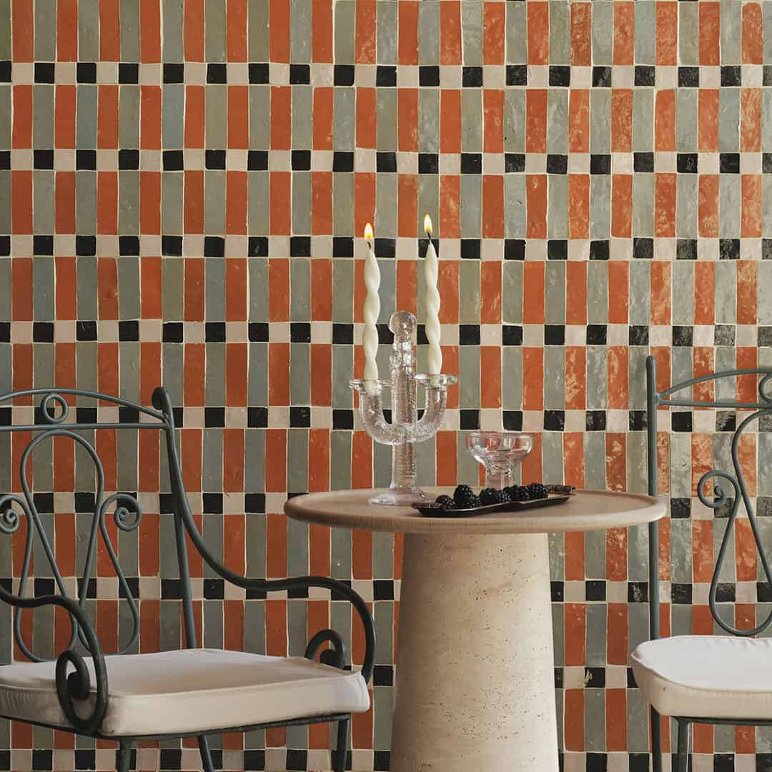 Cozy setting with a round table, vintage chairs, and a textured orange and black tile wall. Candles, a crystal glass, and a small dish of blackberries create an inviting atmosphere.