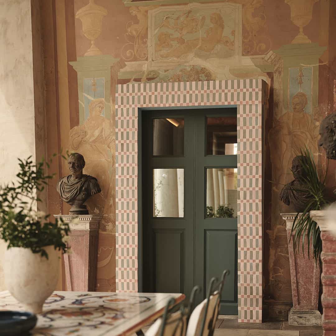 Elegant room featuring a green door framed by pink and white tiles, surrounded by classical busts, frescoed walls, and a rustic table with chairs.