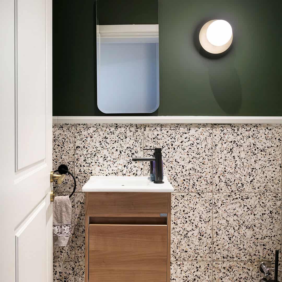 Modern bathroom vanity with terrazzo wall tiles, wood cabinet, black faucet, and green painted wall with round light.