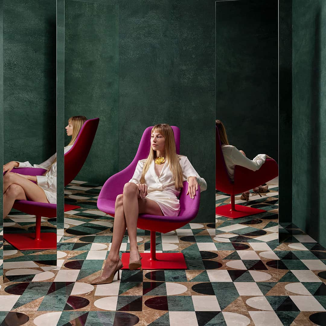 Woman seated in a modern purple chair surrounded by mirrors, with bold geometric patterned tile flooring and deep green walls.