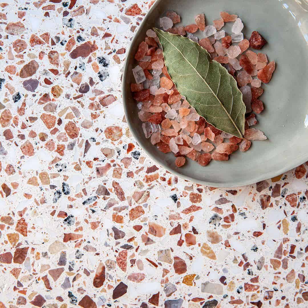 Close-up of terrazzo surface with warm-toned stone chips, featuring a bowl of pink salt and a bay leaf.