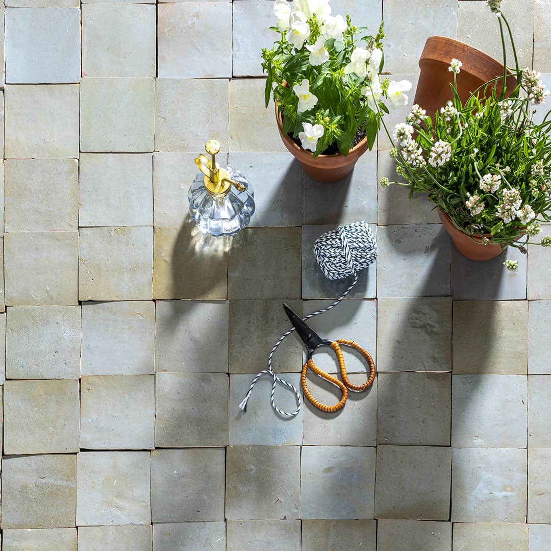 A flat lay on light gray square tiles showing two terracotta pots with white flowering plants, a small glass spray bottle with a gold nozzle, a spool of black-and-white twine, and orange-handled scissors. The arrangement suggests a gardening theme with soft shadows adding depth.