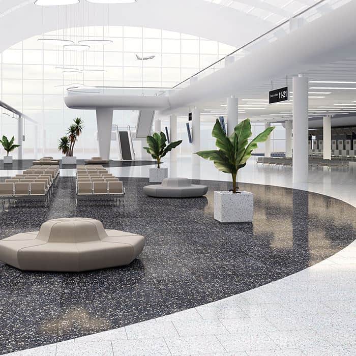 Spacious modern airport terminal with terrazzo flooring, circular beige seating, large glass windows, tropical potted plants, and overhead signage for gates 11–21.