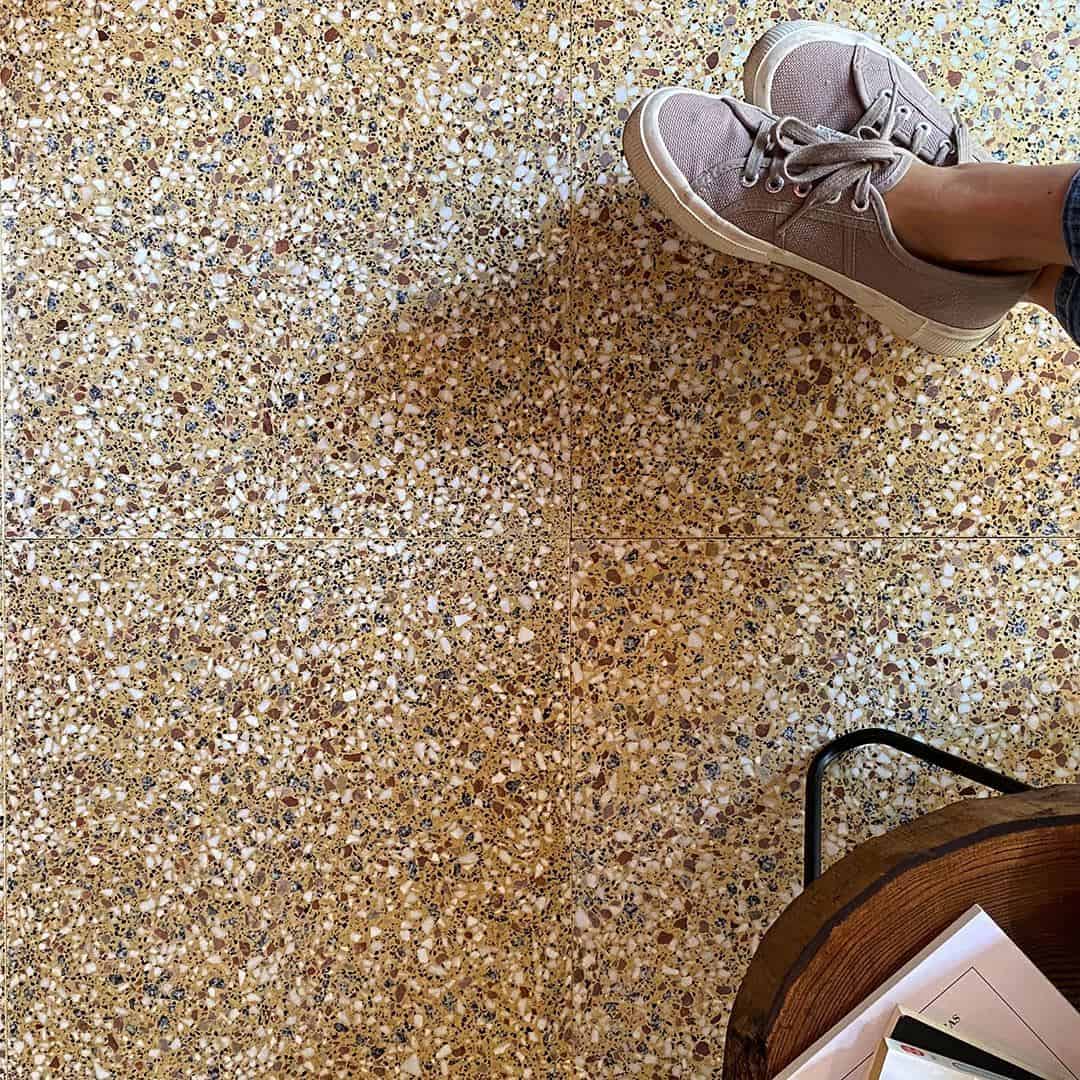 Warm-toned terrazzo tile floor with brown and cream chips, styled with sneakers and wood table edge.