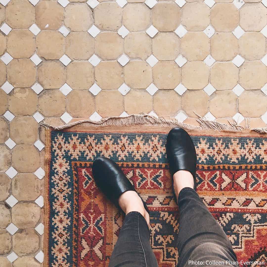 a person wearing black slip-on shoes standing on a colorful geometric rug with red, blue, beige, and orange tones, fringed at the top, placed over tan hexagonal tiles with white diamond inserts.