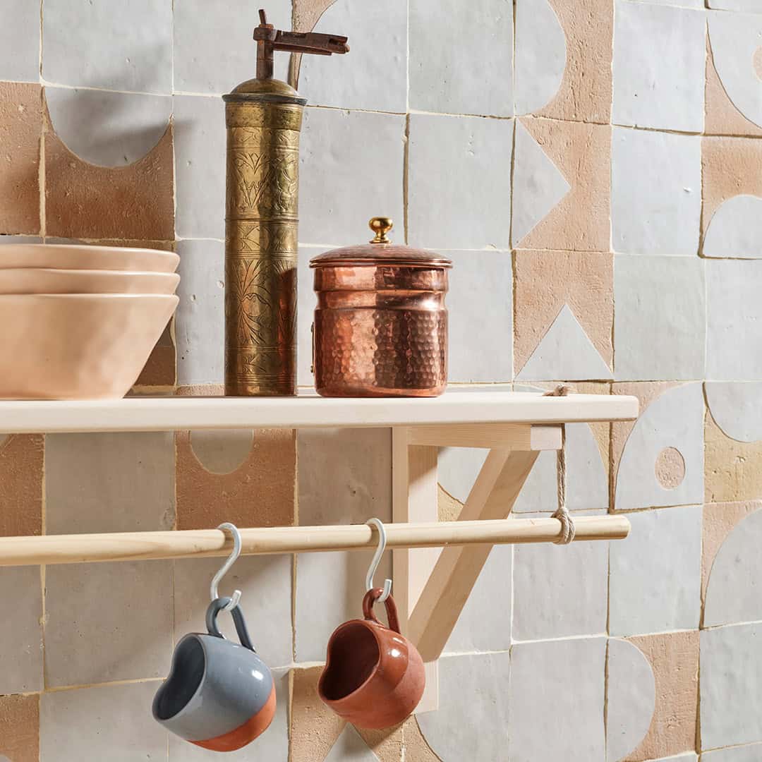 Wooden shelf with glass cups, terracotta bowls, a vintage coffee grinder, and hanging mugs against a geometric tile wall in earthy tones.
