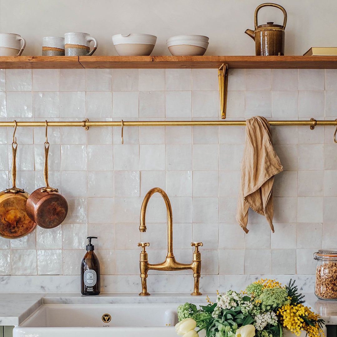 A rustic kitchen scene with a white tiled backsplash, brass faucet, and brass rail holding copper pots and a beige cloth.