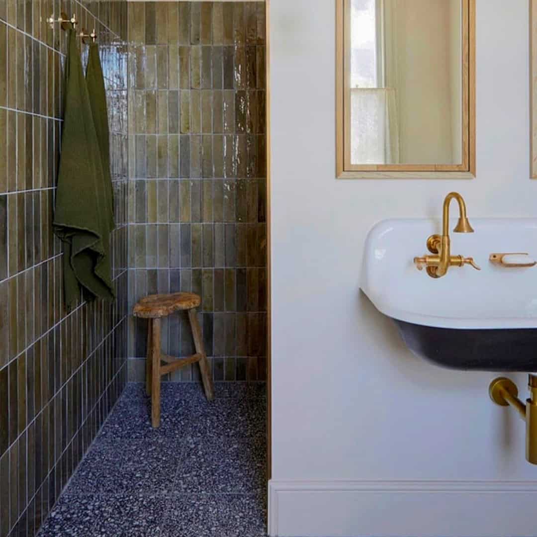 Bathroom with green tiled shower, terrazzo floor, wooden stool, and white sink with brass fixtures.