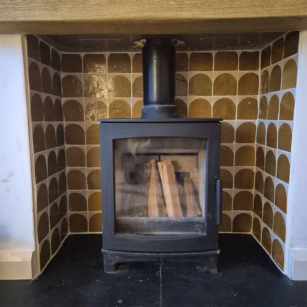 Hand‑made Amber Arch Terracotta Zellige Moroccan tiles with glossy amber and terracotta tones installed as a decorative fireplace surround behind a black wood‑burning stove.