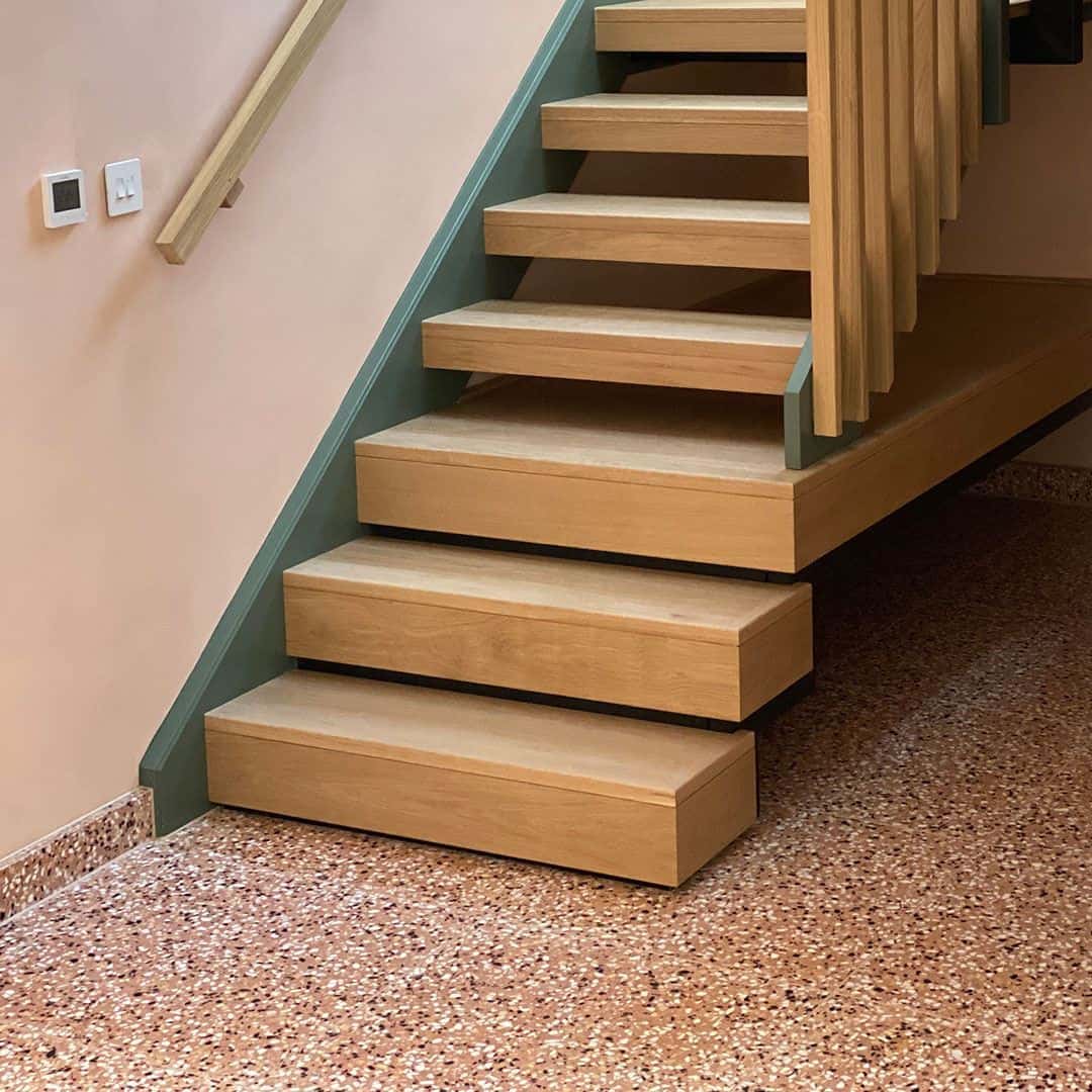 Wooden staircase with open risers above speckled terrazzo flooring and soft neutral-toned walls.