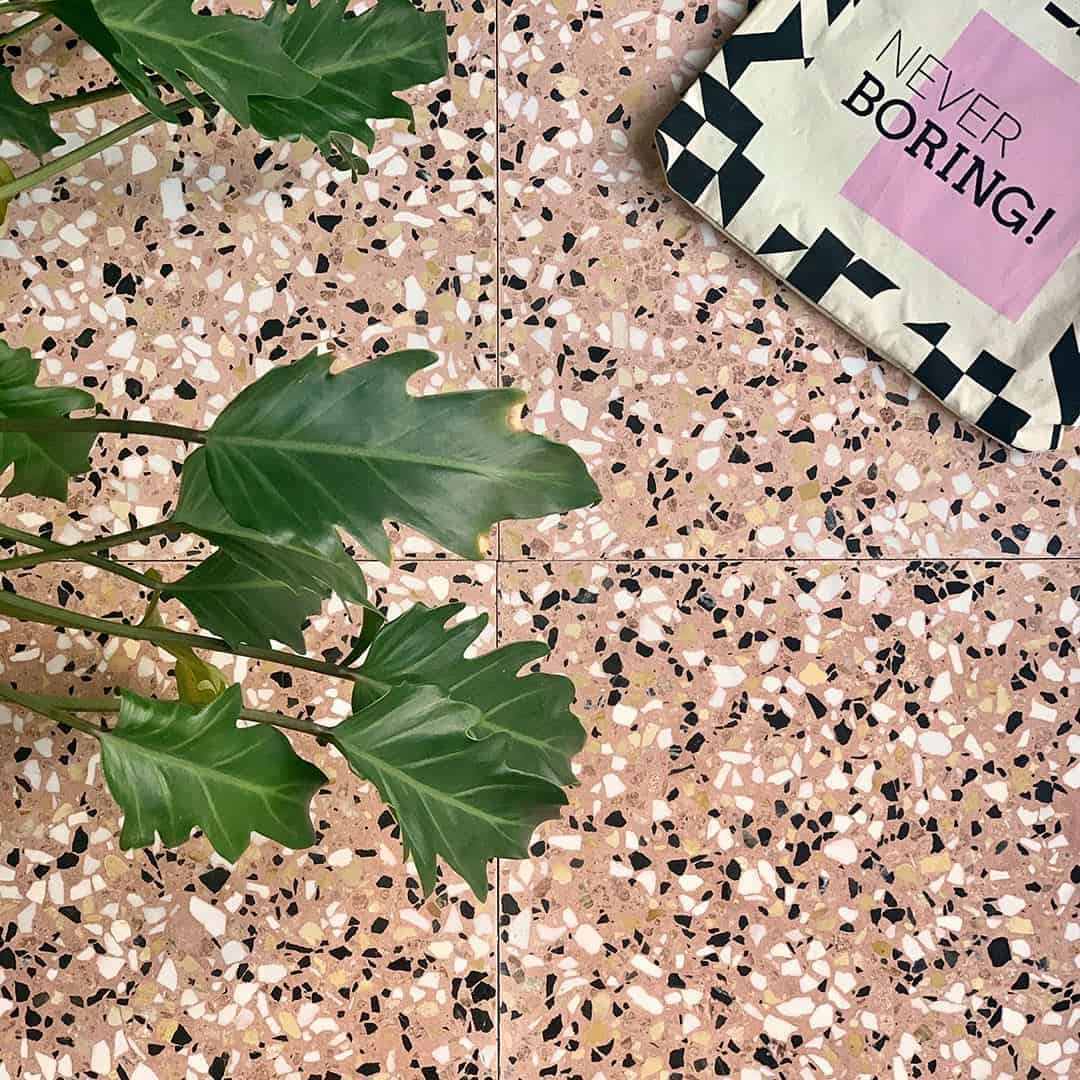Pink terrazzo tile floor with black and white chips, styled with green plant leaves and tote bag.