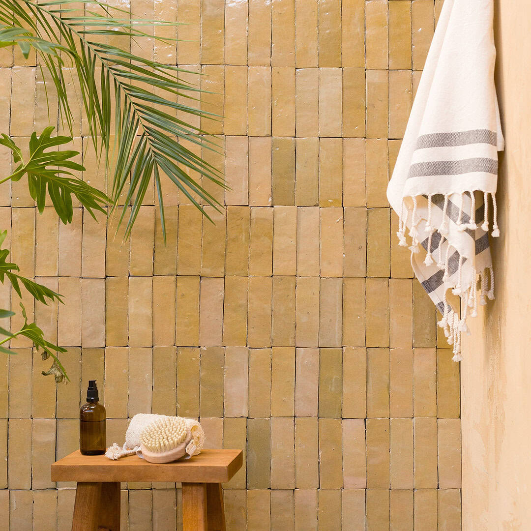 Bathroom scene with beige tiled wall, wooden stool holding brown glass pump bottle and white bath brush, striped towel hanging, and palm leaves adding natural, spa-like calming atmosphere.