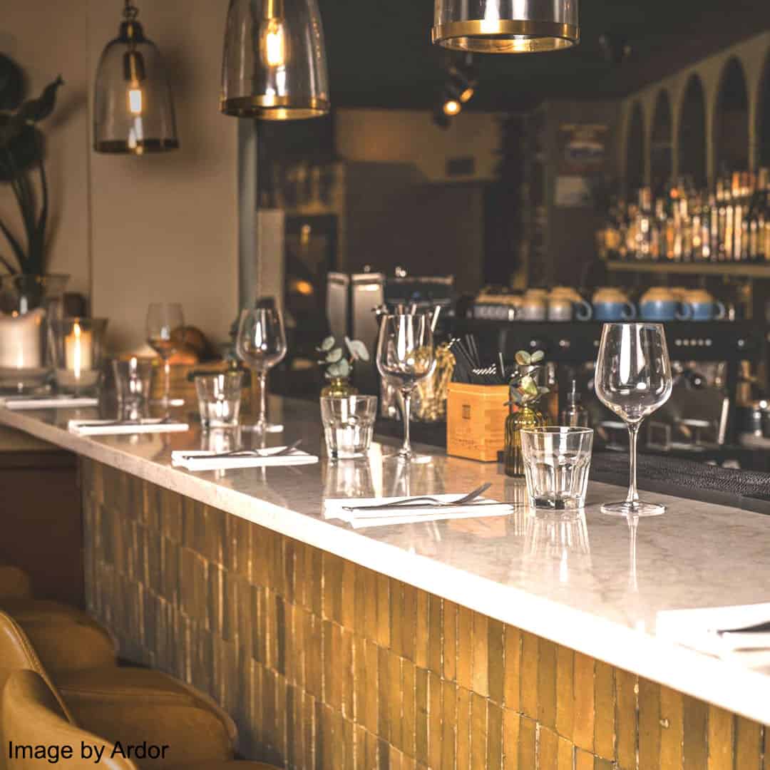 Elegant bar counter with polished marble surface and golden textured front, set with wine glasses, cutlery, napkins, and greenery vases, illuminated by metallic pendant lights in a stylish restaurant.