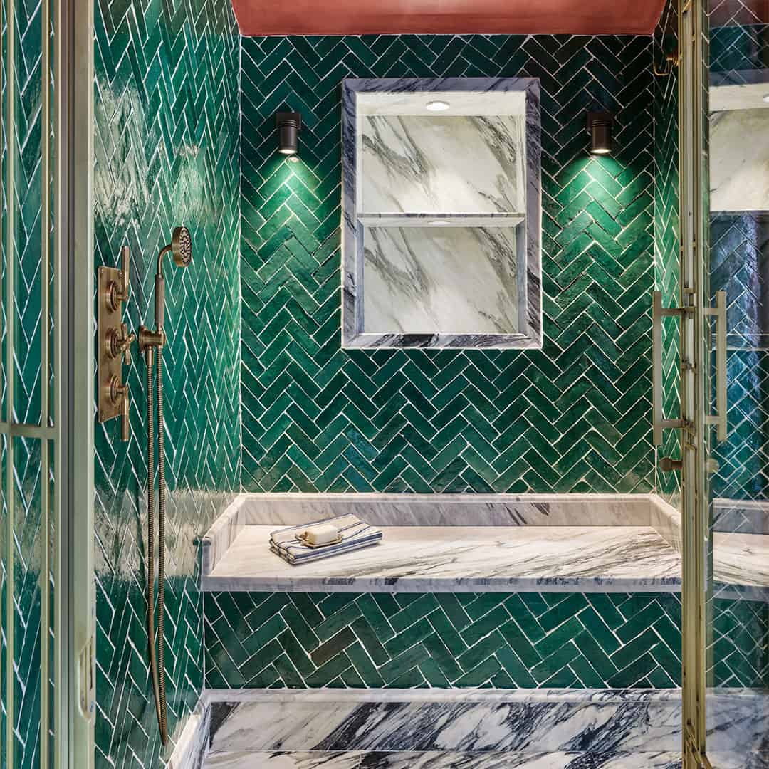 Luxurious shower with glossy green herringbone tiles, marble bench and trim, brass fixtures, handheld showerhead, recessed niche, and wall-mounted lights illuminating elegant, modern bathroom design with rich textures.